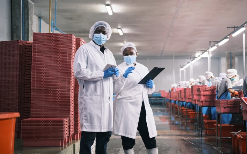 Two food production workers wearing lab coats, hairnets, gloves, and face masks reviewing information on a tablet and clipboard inside a processing plant.
