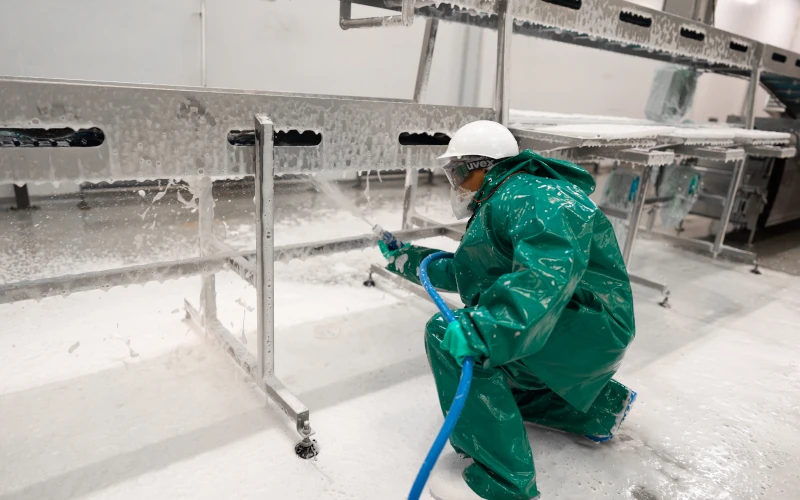 Worker in green protective suit and helmet using a high-pressure hose to clean stainless steel equipment during sanitation in a food production environment.