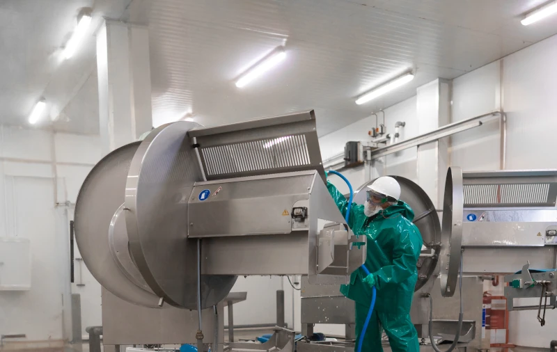 Worker in green protective clothing using a hose to wash down large stainless steel machinery inside a food processing facility.