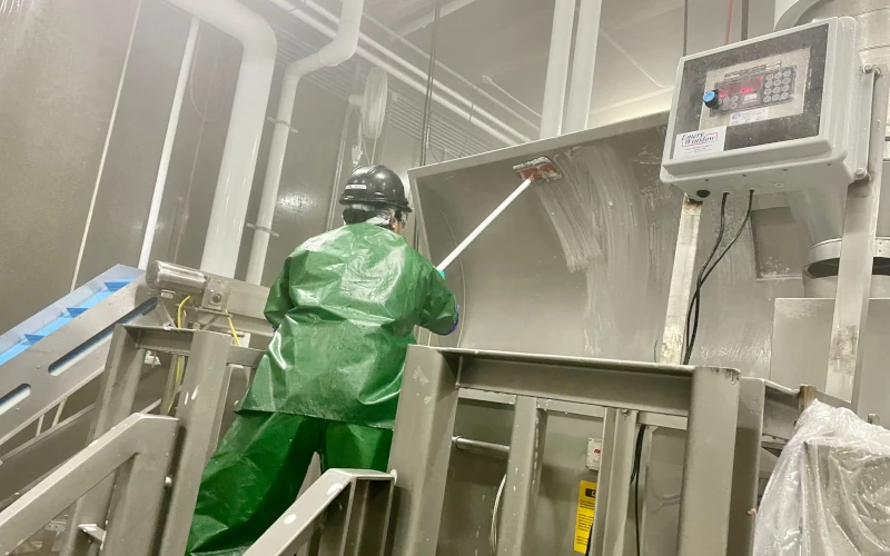Worker in green protective clothing using a long-handled brush to clean large stainless steel machinery during sanitation in a food processing facility.