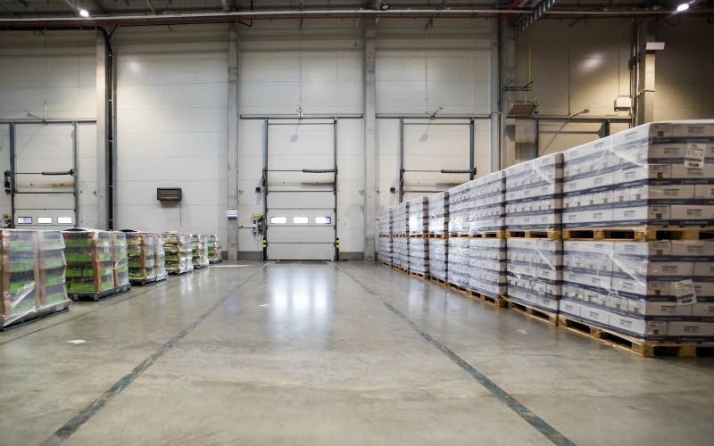 Large industrial warehouse interior with pallets of packaged products stacked and prepared for distribution near loading dock doors.