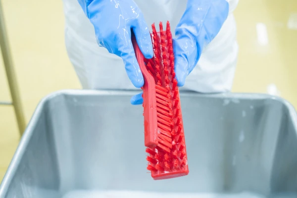 Food processing worker wearing protective gloves and holding a red cleaning brush over a container during sanitation.