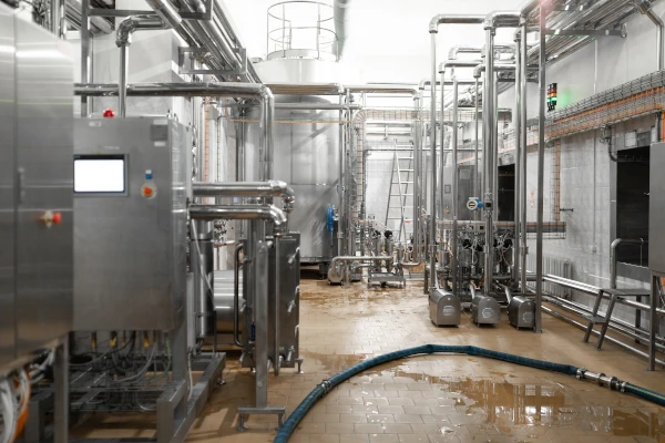 Stainless steel pipes and industrial processing equipment inside a food processing facility with a wet production floor.